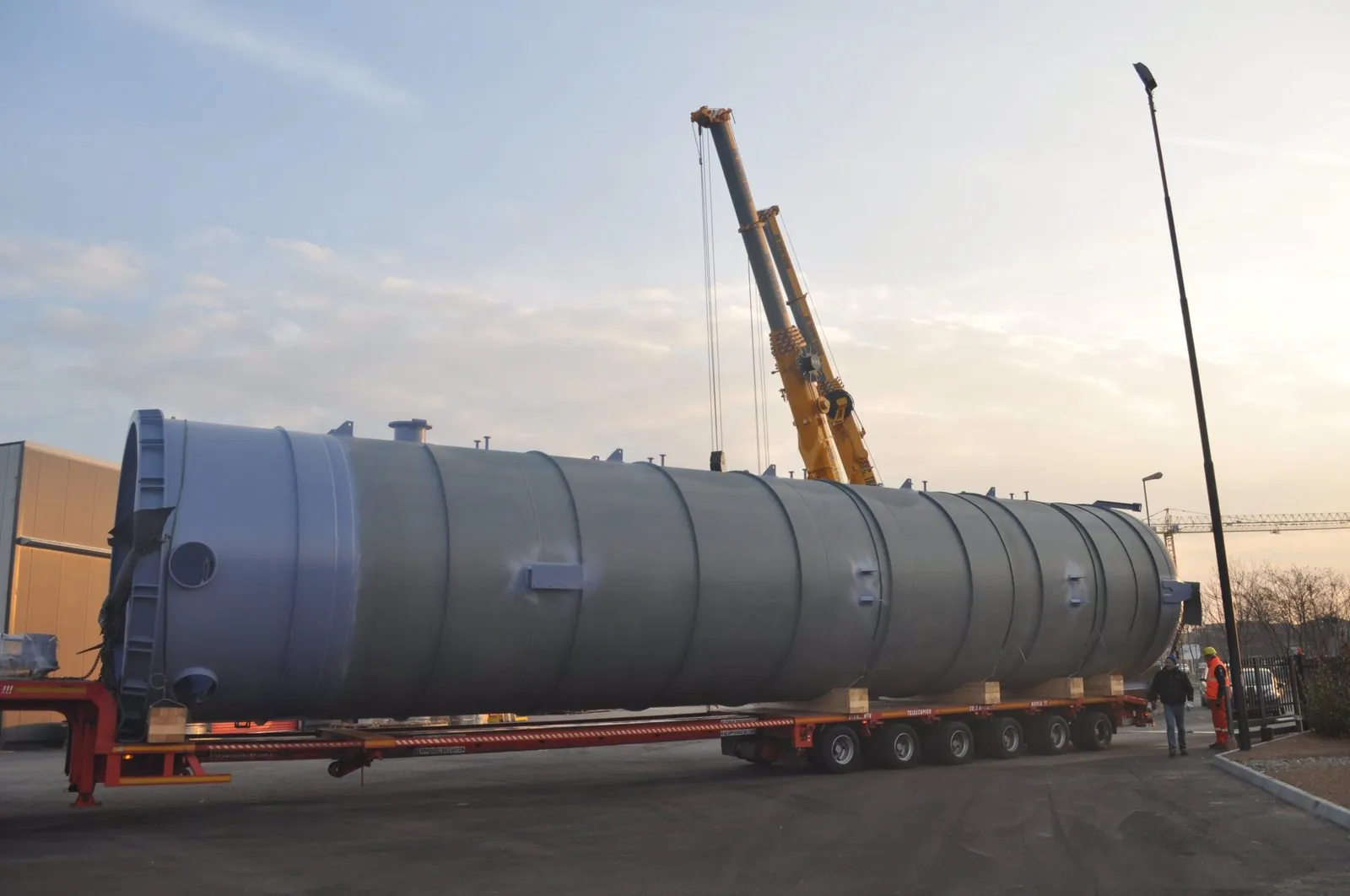 Large water storage tank on a multi-axle low-bed trailer at dusk, ISCOTRANS team alongside